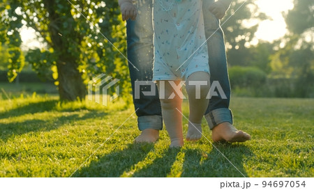 Walking children's bare feet on a green lawn close-up. Child learns to take the first steps on the grass. Baby learns to walk with the help of his mother on a green grass in the park. Walking children's bare feet on a green lawn close-up. Child learns to take the first steps on the grass. Baby learns to walk with the help of his mother on a green grass in the park. 94697054