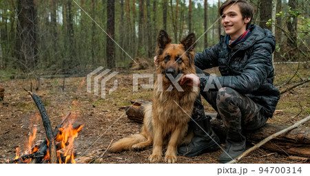Young man with big dog sits at the fire in the forest. Cheerful guy is stroking his dog, outdoor. Happy caucasian teenager with his German shepherd by the fire in nature, in autumn. Young man with big dog sits at the fire in the forest. Cheerful guy is stroking his dog, outdoor. Happy caucasian teenager with his German shepherd by the fire in nature, in autumn. 94700314