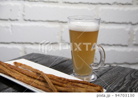 Thin sausages for beer. On a rectangular plate. Next to it is a glass of wheat beer. Background from pine boards. Close-up. Thin sausages for beer. On a rectangular plate. Next to it is a glass of wheat beer. Background from pine boards. Close-up. 94703130