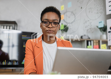 Portrait of african business woman in orange suit sitting at desk and looking at camera. Portrait of african business woman in orange suit sitting at desk and looking at camera. 94704003