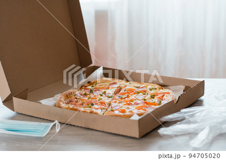 Takeaway food. Pizza in a cardboard box, gloves and protecrive mask on the table in the kitchen. Takeaway food. Pizza in a cardboard box, gloves and protecrive mask on the table in the kitchen. 94705020