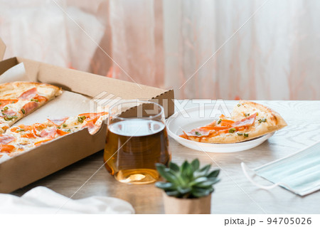 Takeout food. A slice of pizza in a disposable plastic plate and a box of pizza on the table in the kitchen. 94705026
