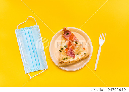 Takeaway and delivery. A slice of pizza in a disposable plastic plate, a protective mask and a fork on a yellow background Takeaway and delivery. A slice of pizza in a disposable plastic plate, a protective mask and a fork on a yellow background 94705039