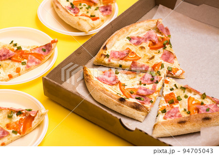 Takeaway and delivery. Pizza in a cardboard box and pieces of pizza laid out on disposable plates on a yellow background Takeaway and delivery. Pizza in a cardboard box and pieces of pizza laid out on disposable plates on a yellow background 94705043