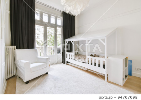 Interior of white room with baby crib and armchair near window with blue chest of drawers in daylight 94707098