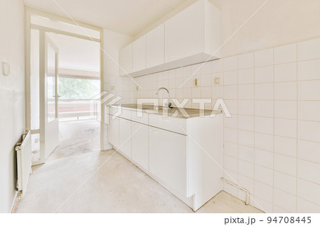 Fragment of interior of light narrow home kitchen with minimalist style white furniture with sink and stove Fragment of interior of light narrow home kitchen with minimalist style white furniture with sink and stove 94708445