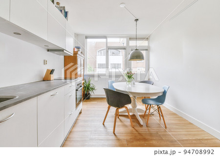 Modern minimalist style interior design of studio apartment with open white kitchen and dining zone with table and chairs illuminated by loft style lamp 94708992