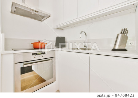 Fragment of interior of light narrow home kitchen with minimalist style white furniture with sink and stove 94709423