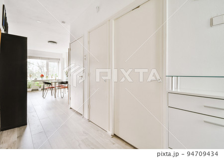 Narrow corridor with white walls and doors leading to spacious room with windows and parquet floor in modern apartment 94709434