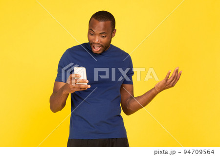 Discontented Black Male Athlete Holding Phone Over Yellow Studio Background Discontented Black Male Athlete Holding Phone Over Yellow Studio Background 94709564