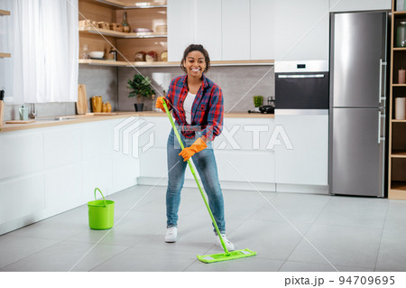 Happy young black woman in rubber gloves washes floor with mop in minimalist kitchen interior, full length 94709695