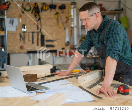 Portrait of a carpenter in protective glasses and work overalls uses a laptop in a workshop. Portrait of a carpenter in protective glasses and work overalls uses a laptop in a workshop. 94711210