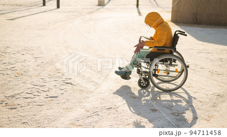 Caucasian woman in a wheelchair uses a smartphone on a walk in the park in winter. Caucasian woman in a wheelchair uses a smartphone on a walk in the park in winter. 94711458