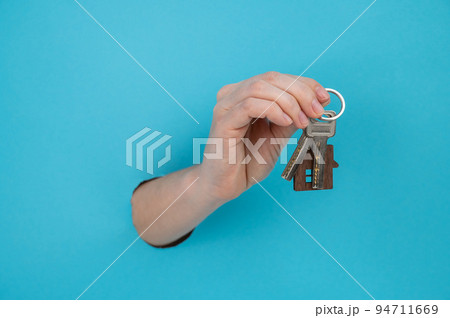 A woman's hand holds the keys with a keychain in the shape of a house on a blue background.  94711669