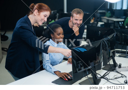 Colleagues look at the monitor and decide working moments. African young woman, caucasian man and red-haired caucasian woman communicate in the office.  94711753