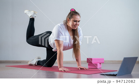 Young woman doing sports exercises at home. The girl is engaged in fitness at home on the mat and watches a training video on a laptop 94712082