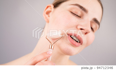 Close-up portrait of a woman using a quartz roller massager on her cheek for an alternative anti-aging. 94712346