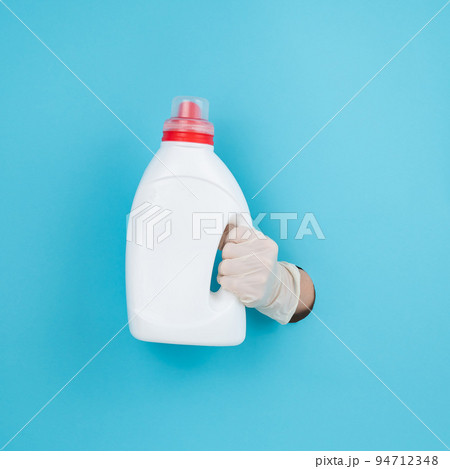 A woman is holding a white bottle of detergent. Woman's hand sticking out of blue paper background. A woman is holding a white bottle of detergent. Woman's hand sticking out of blue paper background. 94712348