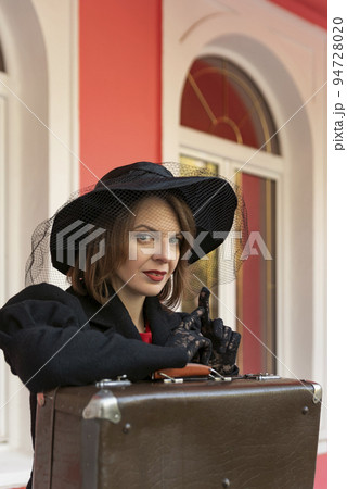 Portrait of beautiful aristocratic woman in black wide-brimmed hat and lace gloves with retro suitcase. Female traveler 94728020
