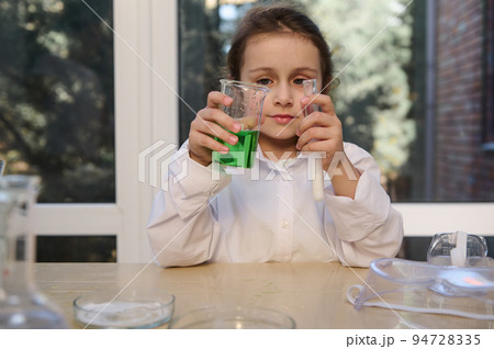 Details: hands of a little girl, a chemist in white lab coat, hold a test tube and a flask, carry out a chemical reaction, pour a solution of soda into citric acid, observe the chemical reaction 94728335