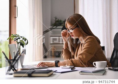 Young businesswoman holding her glasses and looking at laptop screen, working in internet at her office desk 94731744