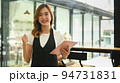 Smiling coffee shop owner wearing apron using digital tablet while standing at her coffee shop 94731831