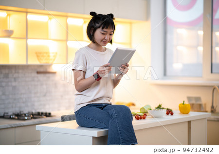 Young woman with a gadget sitting in the kitchen Young woman with a gadget sitting in the kitchen 94732492
