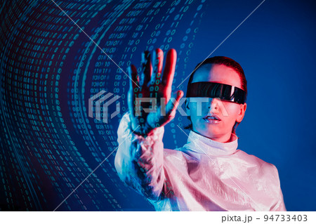 Woman dressed in holographic clothes and futuristic eyeglasses with reflection of matrix code is touching by hand matrix digital numbers artificial intelligence data before her in blue light 94733403