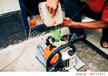 Refilling chainsaw tank with fuel gasoline mixture. Closeup view on hand selective focus. Electric tools maintenance for gardening work and lawn mowing concept Refilling chainsaw tank with fuel gasoline mixture. Closeup view on hand selective focus. Electric tools maintenance for gardening work and lawn mowing concept 94737621