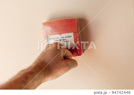 Male fist breaks plastic cover of red fire alarm button 94742446