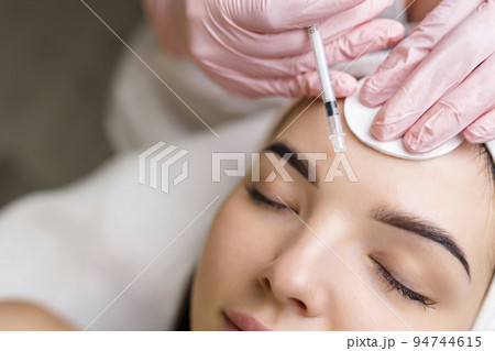 Close up of hands of young cosmetologist injecting botox in female face. She is standing and smiling. The woman is closed her eyes with relaxation 94744615