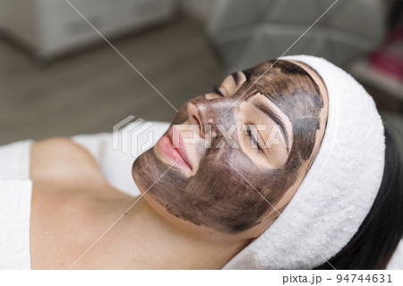 cosmetologist applying black mask on pretty woman face wearing black gloves, gorgeous woman in spa having facial procedures 94744631