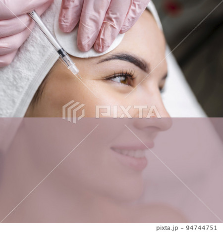 Close up of hands of young cosmetologist injecting botox in female face. She is standing and smiling. The woman is closed her eyes with relaxation 94744751