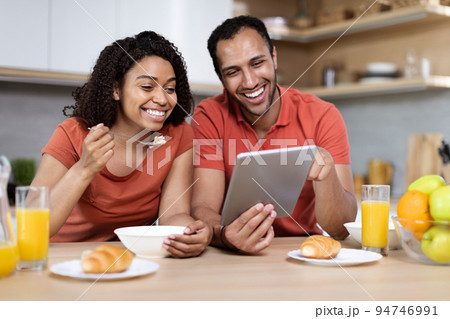 Laughing millennial black lady and guy enjoy breakfast in morning, watch video on tablet together in kitchen interior 94746991