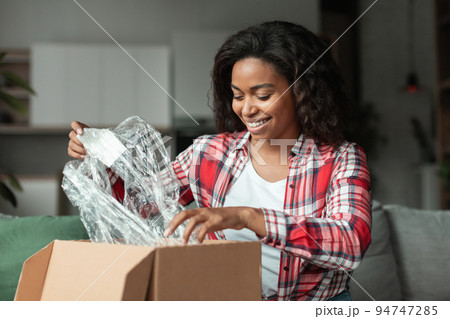 Cheerful millennial african american woman opens cardboard box and unpacks parcel in living room 94747285