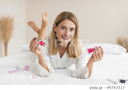 Happy mature woman in white robe lying on bed and choosing nail polish, making manicure in bedroom interior 94747317