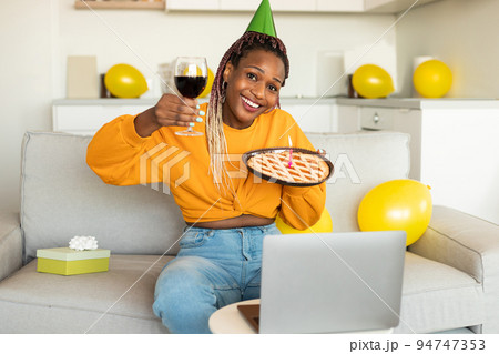Online birthday. Excited african american woman celebrating b-day showing pie and glass of wine, video calling on laptop 94747353