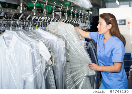 Adult female worker of laundry inspecting clothing after cleaning on racks 94748180
