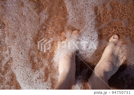 Top view of men's legs in the sea. A man is standing on the sand. 94750731