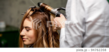 The female hairdresser is curling hair for a brown-haired young caucasian woman in a beauty salon 94762208
