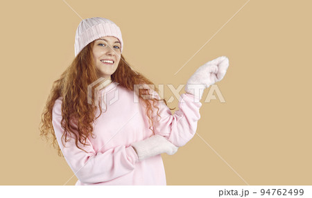 Happy woman in pink winter hat and mittens points hand to side on beige background 94762499