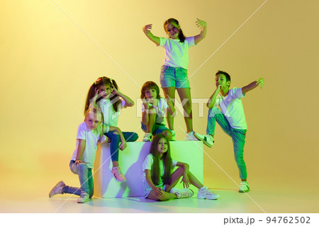 Dance class. Joyful children, kids in t-shirts and jeans dancing in action isolated on green background in yellow neon light. Dance class. Joyful children, kids in t-shirts and jeans dancing in action isolated on green background in yellow neon light. 94762502