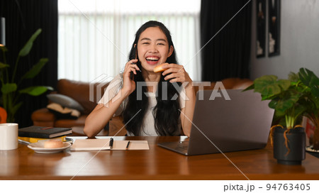 Smiling young asian woman sitting front of laptop at her home office and talking on mobile phone Smiling young asian woman sitting front of laptop at her home office and talking on mobile phone 94763405
