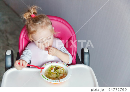 Infant baby with dirty face eating soup herself with spoon. Cute little girl sitting in high baby chair in kitchen at home. Maternity family childhood concept 94772769