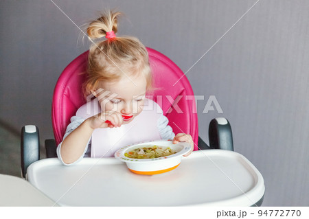 Infant baby with dirty face eating soup herself with spoon. Cute little girl sitting in high baby chair in kitchen at home. Maternity family childhood concept Infant baby with dirty face eating soup herself with spoon. Cute little girl sitting in high baby chair in kitchen at home. Maternity family childhood concept 94772770