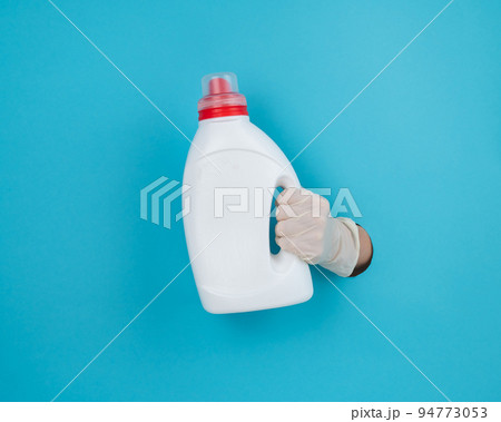 A woman is holding a white bottle of detergent. Woman's hand sticking out of blue paper background. A woman is holding a white bottle of detergent. Woman's hand sticking out of blue paper background. 94773053
