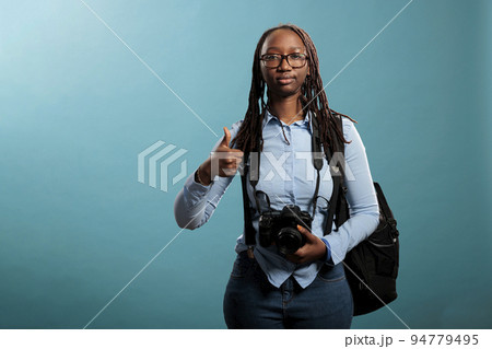 Joyful photographer giving ok finger gesture while standing on blue background. Cheerful professional photography entusiast with DSLR device saying is ready for photo shooting by giving thumbs up sign Joyful photographer giving ok finger gesture while standing on blue background. Cheerful professional photography entusiast with DSLR device saying is ready for photo shooting by giving thumbs up sign 94779495