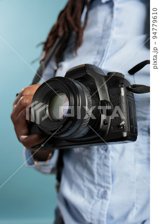 Close up of young adult photograhpy entusiast holding a modern camera device while standing on blue background. Professional photographer with DSLR enjoying leisure time. Studio shot Close up of young adult photograhpy entusiast holding a modern camera device while standing on blue background. Professional photographer with DSLR enjoying leisure time. Studio shot 94779610