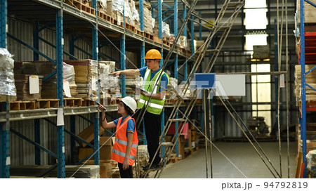 Male and female workers wearing hardhats and reflective jackets checking inventory boxes on shelf with barcode scanner 94792819