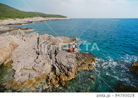 Man and woman are standing on a rocky cliff near the sea against the backdrop of mountains Man and woman are standing on a rocky cliff near the sea against the backdrop of mountains 94793028